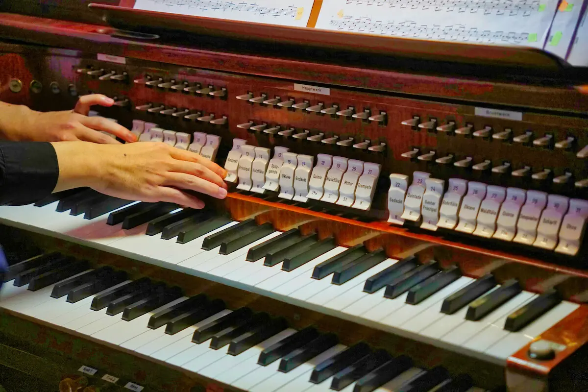 Blick auf die Klaviatur einer Orgel. Links im Bild die Hände des Organisten beim Bedienen der Register. Oberhalb der Register sind Notenblätter im Bild.