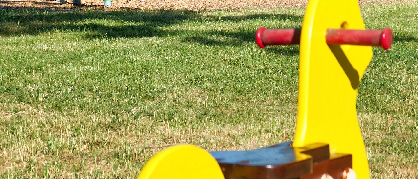 Blick auf einen Kinderspielplatz mit diversen Spielgeräten.  Im Bildvordergrund ist ein Schaukelpferd zu sehen. Im Hintergrund sind Klettergerüste und eine Rutschbahn im Bild.