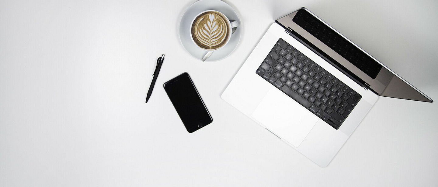 Arbeitstisch von oben mit Laptop, Kaffeetasse, Smartphone und Stift.