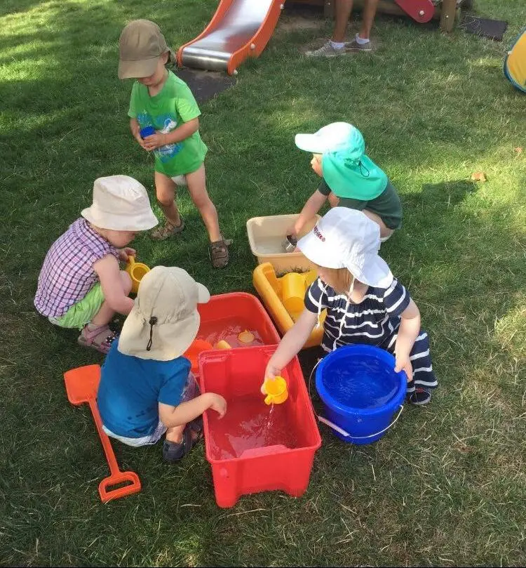 Das Bild zeigt eine Gruppe von fünf kleinen Kindern, die auf einer Rasenfläche spielen. Sie tragen Hüte zum Schutz vor der Sonne und sind mit verschiedenen Spielsachen beschäftigt, darunter Eimer, Schaufeln und Wasserbehälter. Einige Kinder scheinen mit Wasser zu spielen. Im Hintergrund ist eine rote Rutsche sichtbar. Die Szene wirkt fröhlich und aktiv.