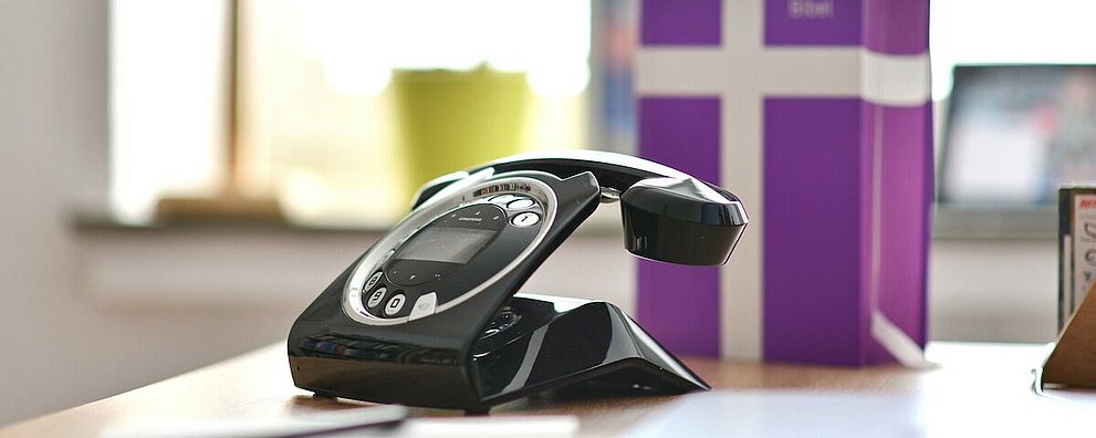 Schreibtisch mit Focus auf Telefon, die Basisbibel unscharf dahinter. Im Hintergrund in der Unschärfe, die Fensterbank mit Kreuz und Blumentopf.