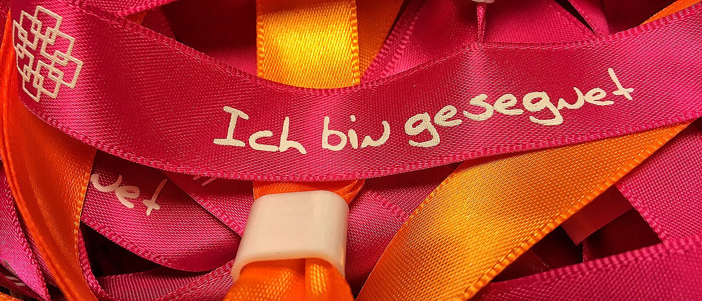 Rote und orangene Stoffarmbänder mit der Aufschrift "Ich bin gesegnet" und dem Facettenkreuz.
