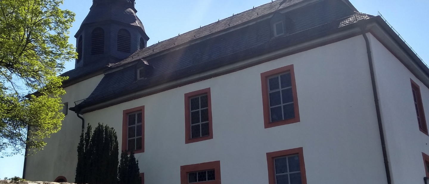 Weißes Kirchengebäude mit dem Turm links und einem Baum