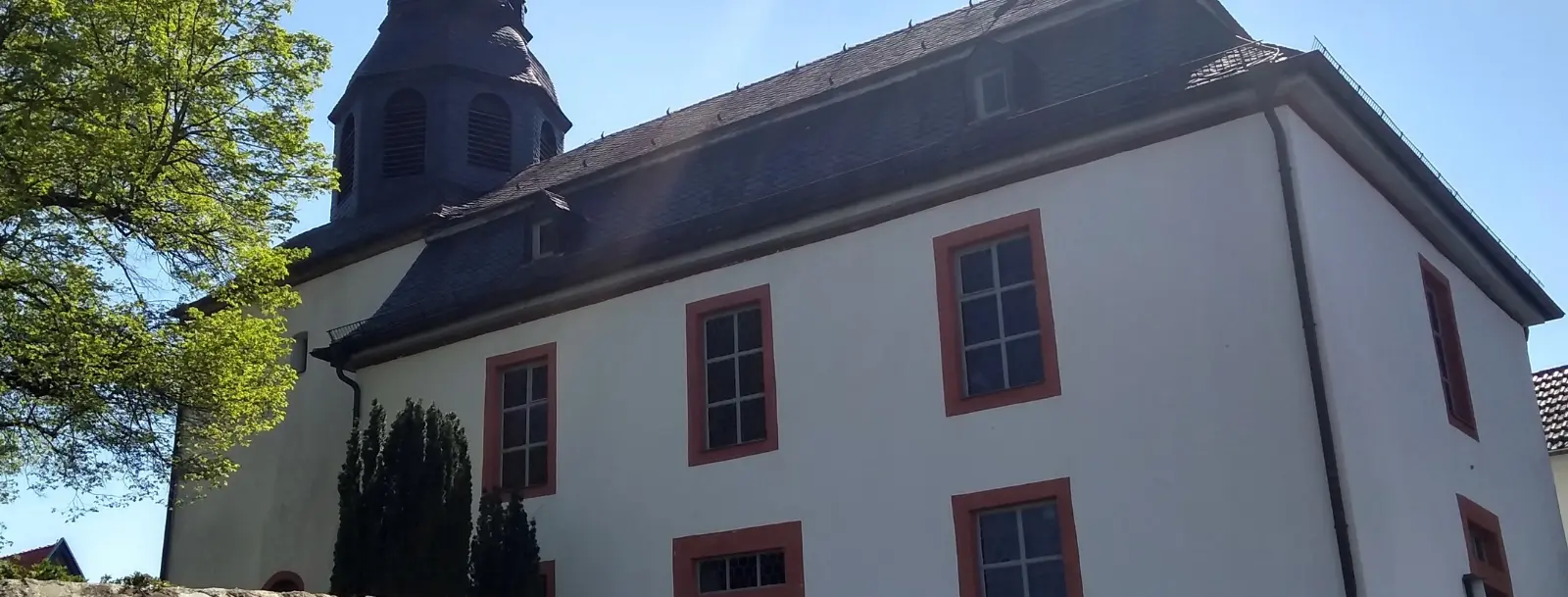 Weißes Kirchengebäude mit dem Turm links und einem Baum