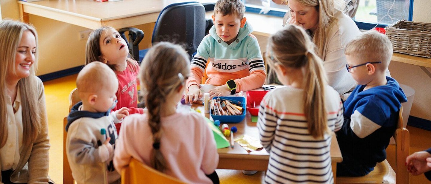 Ev. Kindertagesstätte "Kinder Garten Eden" Gemünden. Blick auf einen Tisch mit spielenden Kindern. Es sitzt noch eine Erzieherin mit am Tisch. Eine weitere Erzieherin kniet auf dem Teppich.