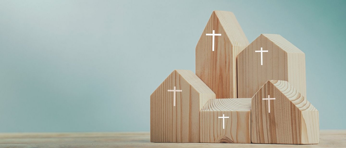 Kleine Häuschen aus Holzblöcken mit einem weißen Kreuz stehen als Kirchensymbole in einer Gruppe in der rechten Bildhälfte. Der Hintergrund ist einfach blaugrün 
