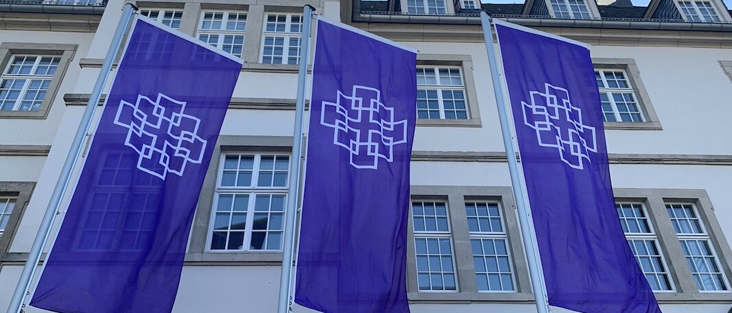 Drei lila Flaggen mit dem Facettenkreuz vor einem Gebäude