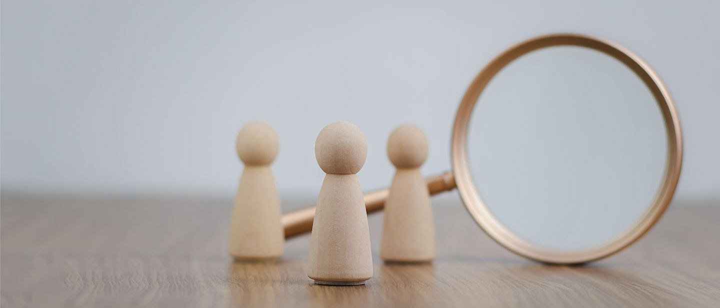 drei kleine Holzkegelfiguren stehen auf einer Tischplatte. Dahinter steht aufrecht auf der Rundung ruhend eine Lupe