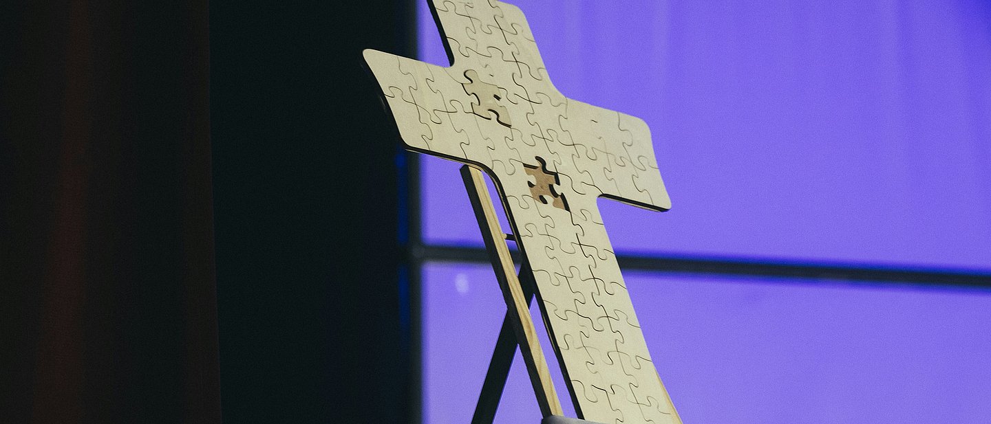 Das Foto zeigt ein Kreuz auf einem Altar, das als Puzzle dargestellt wird, in dem ein Puzzle-Teil fehlt