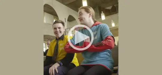 Zwei junge Menschen sitzen nebeneinander in einer Kirche und lächeln. Über dem Bild liegt ein Abspiel-Symbol, das auf ein Video hinweist.