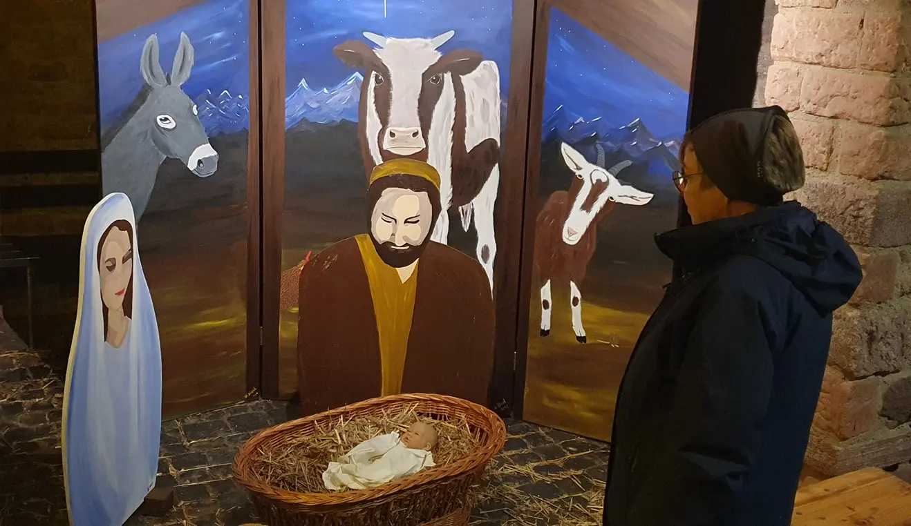 Eine Person betrachtet große Figuren von Maria und Joseph. Die schauen auf einen kleinen Korb aus Bast mit einer Puppe als Jesuskind. Im Hintgrund große Bilder von Ochs und Esel.