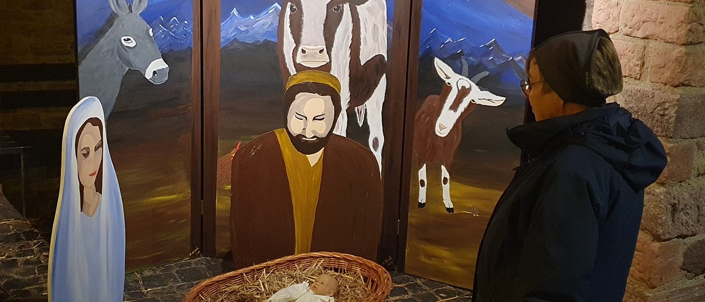 Eine Person betrachtet große Figuren von Maria und Joseph. Die schauen auf einen kleinen Korb aus Bast mit einer Puppe als Jesuskind. Im Hintgrund große Bilder von Ochs und Esel.