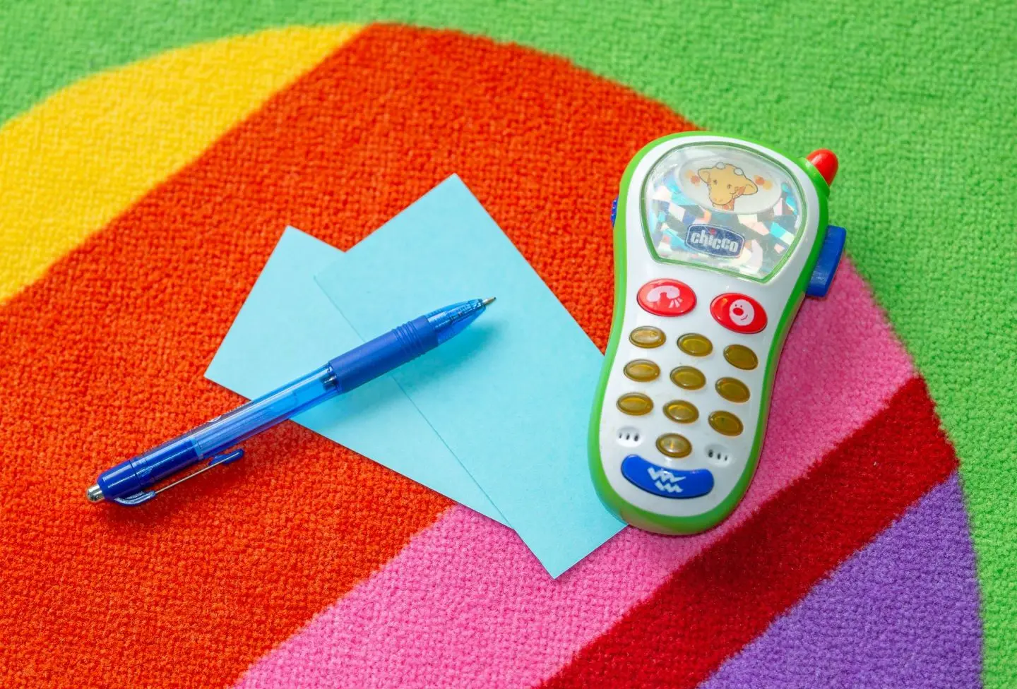 Auf einem bunten Teppich liegen ein Kugelschreiber, zwei blaue Zettel und ein Spielzeugtelefon 