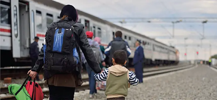 Blick auf eine Mutter, mit einem kleinen Jungen an der Hand, die mit Rucksäcken bepackt über Bahngleise in Richtung eines stehenden Zugs läuft.