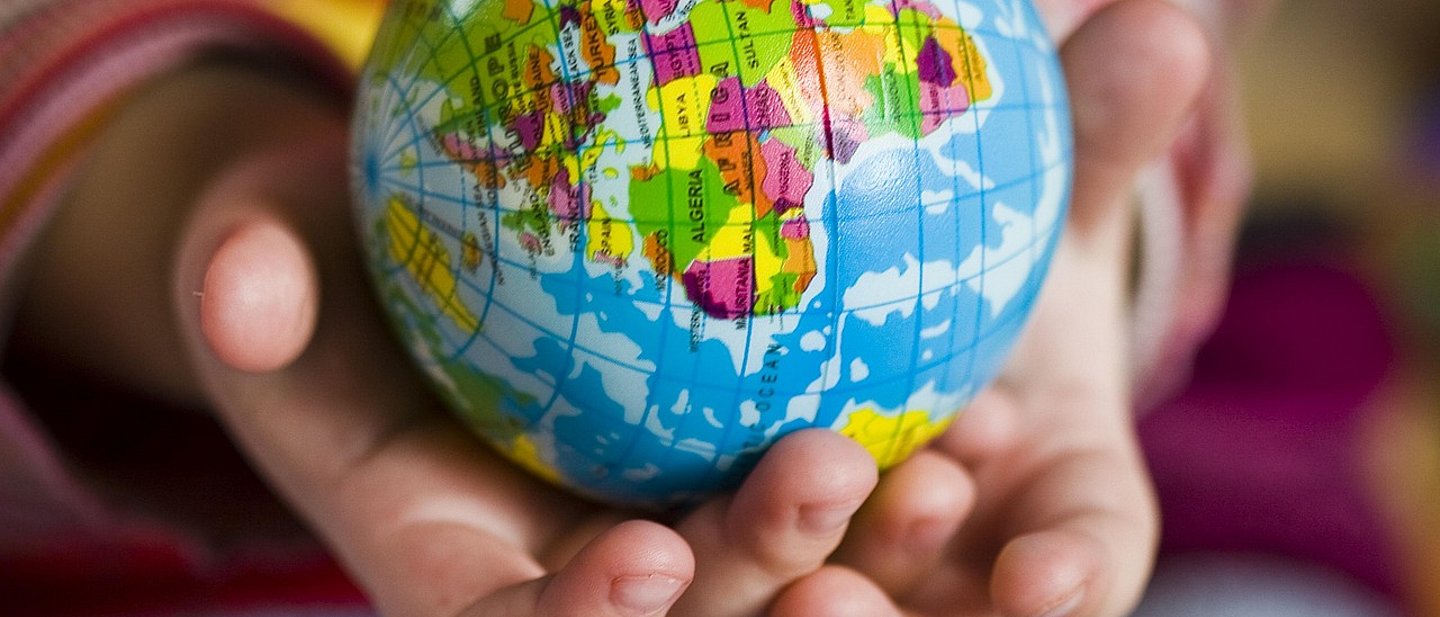 Zwei geöffnete Hände halten einen kleinen Globus mit farbig markierten Ländern. Der Globus zeigt Afrika, Europa und Teile von Asien in kräftigen Farben. Die Hände gehören vermutlich einem Kind, das den Ball behutsam umfasst. Das Bild symbolisiert Fürsorge und weltweite Verbundenheit.