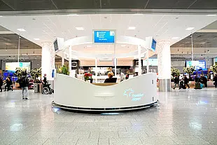 Runder Informations- oder Serviceschalter in der Mitte eines modernen Flughafenterminals. Menschen bewegen sich durch die helle Halle, im Hintergrund sind Geschäfte, Sitzbereiche und digitale Anzeigetafeln zu sehen.