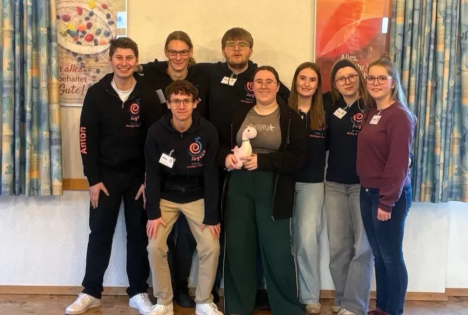 Der 8-köpfige EJVD Vorstand 2026 posiert in einem Gemeindehaus zwischen zwei grünen Vorhängen und einer bunten Wand. 