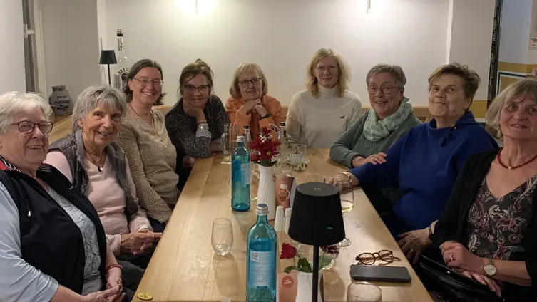 Eine Gruppe von neun älteren Frauen sitzt an einem langen Tisch. Sie schauen alle lächelnd in die Kamera. Auf dem Tisch befinden sich Wasserflaschen und ein paar Dekorationen. Der Raum scheint ein Gemeinschaftsraum zu sein.