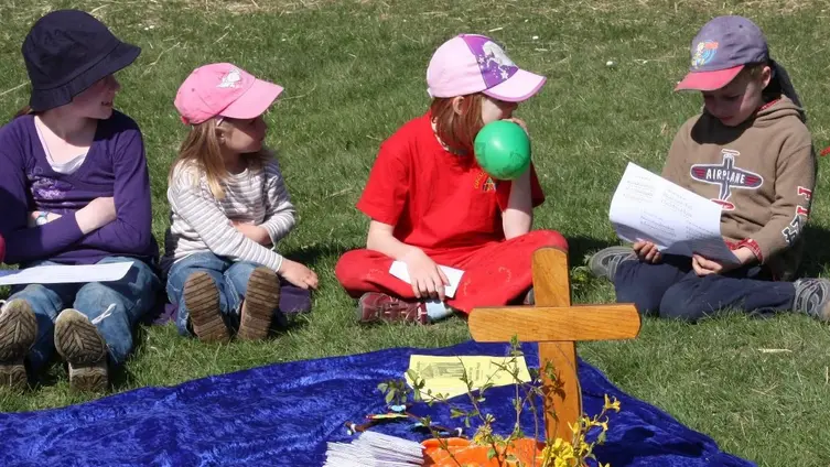 Kinder sitzen beim Familiengottesdienst im Freien rund um eine Altardecke mit Kreuz und Schmuck