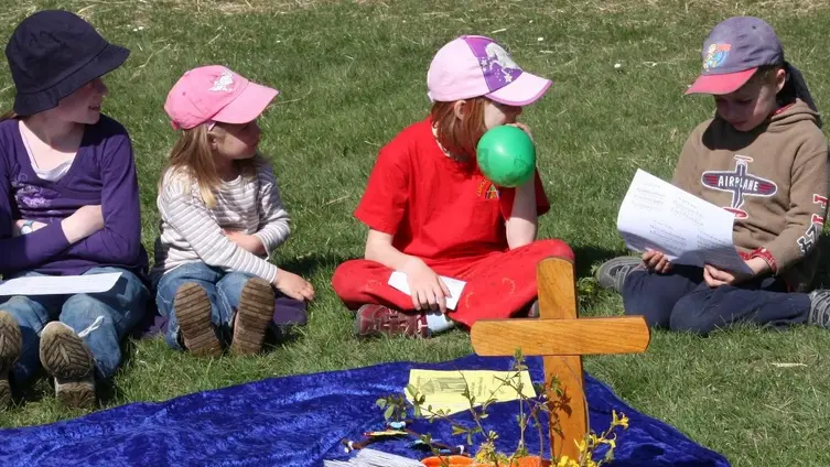 Kinder sitzen beim Familiengottesdienst im Freien rund um eine Altardecke mit Kreuz und Schmuck