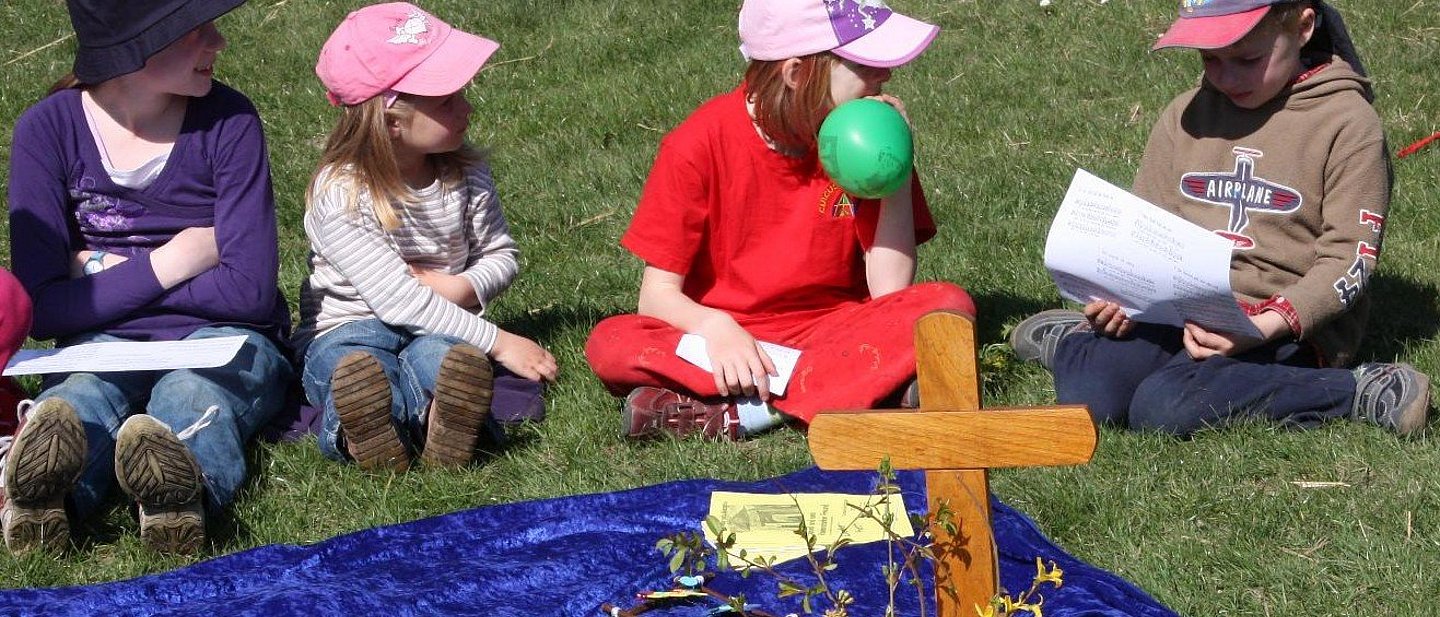 Kinder sitzen beim Familiengottesdienst im Freien rund um eine Altardecke mit Kreuz und Schmuck