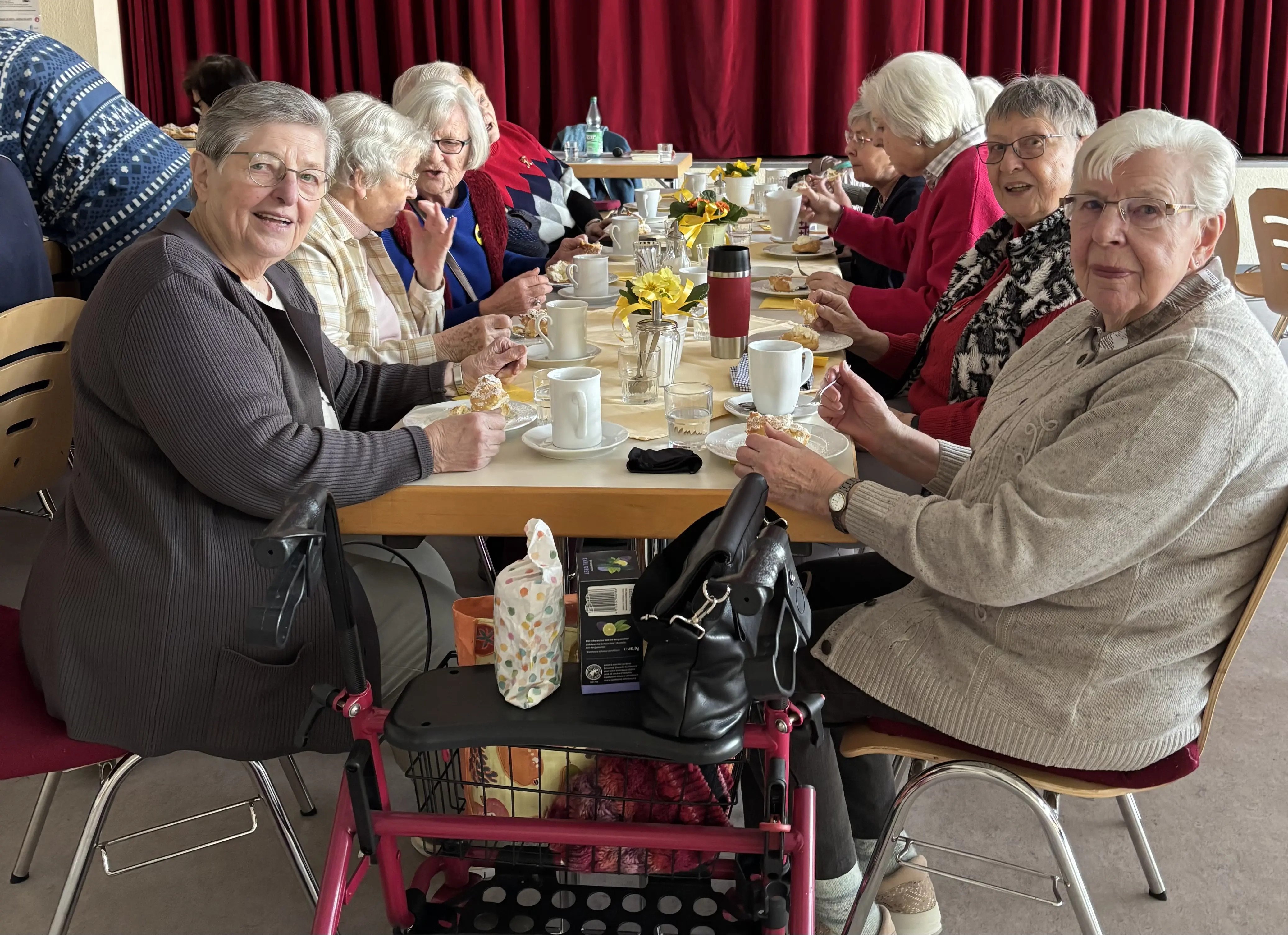 Das Bild zeigt eine Gruppe älterer Damen, die an einem Tisch sitzen und gemeinsam Kaffee trinken oder eine Mahlzeit einnehmen. Sie scheinen sich in einer geselligen Runde zu befinden. Im Hintergrund ist ein roter Vorhang zu sehen, was darauf hindeutet, dass sie sich möglicherweise in einem Gemeinschaftsraum oder einer ähnlichen Einrichtung befinden. Eine der Damen hat einen Rollator vor sich stehen. Die Stimmung wirkt freundlich und entspannt.