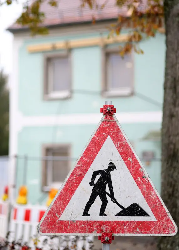 Verkehrsschild Baustelle dreieckig mit rotem Rahmen. Im Hintergrund unscharf das Pfarrhaus