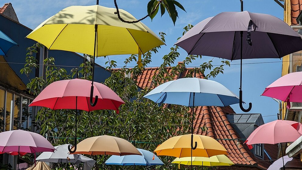 Über der Straße sind viele bunte aufgespannte Regenschirme aufgehängt. Außerdem sind im Bild Hausdächer und das Blattwerk der Bäume zu sehen vor blauem Himmel.