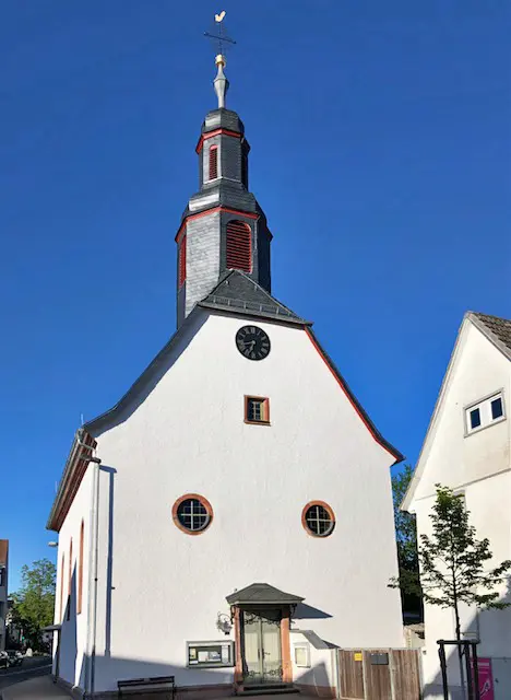 Dorfbarockkirche Köppern, Straßenansicht mit Haupteingang und verschiefertem zweistöckigem Turm. In der Fassade rechts und links über dem Eingang zwei runde Fenster, darüber mittig ein rechteckiges Fenster, darüber die Uhr.