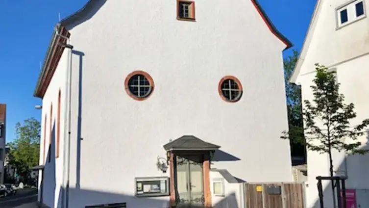 Dorfbarockkirche Köppern, Straßenansicht mit Haupteingang und verschiefertem zweistöckigem Turm. In der Fassade rechts und links über dem Eingang zwei runde Fenster, darüber mittig ein rechteckiges Fenster, darüber die Uhr.