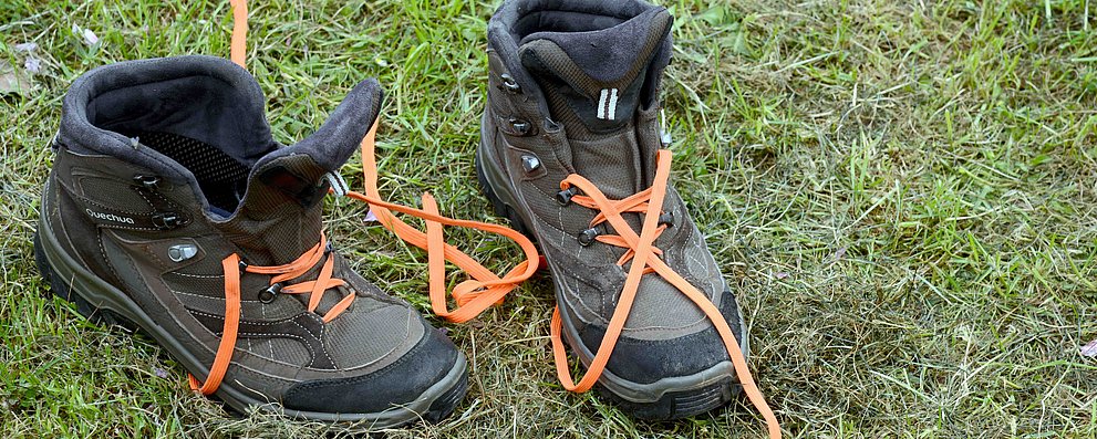 Ein Paar braune Wanderschuhe stehen auf einer Wiese. Die orangefarbenen Schnürsenkel liegen im Gras.