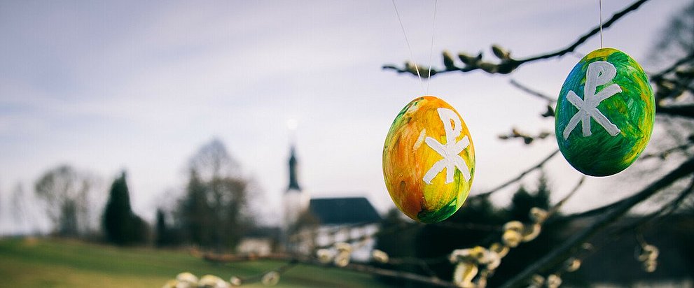 Bunt bemalte Ostereier hängen an einem Baum. Im Hintergrund ist eine Dorfkirche zu sehen.