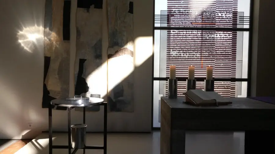 Im Altarbereich steht ein Taufbecken mit Metalloberfläche. Die Sonne scheint darauf und wird reflektiert.