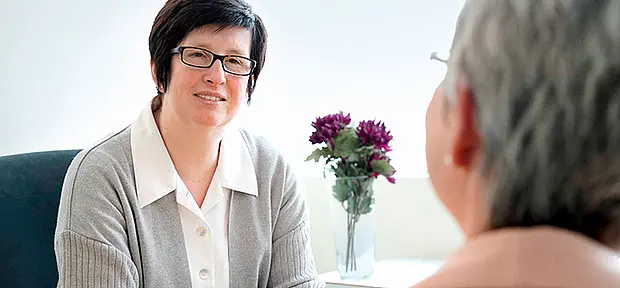 Beratungsgespräch zwischen zwei Frauen in einem hellen Raum. Eine Frau mit Brille sitzt lächelnd an einem Tisch und spricht mit einer älteren Frau, die im Vordergrund von hinten zu sehen ist. Auf dem Tisch steht eine Vase mit Blumen.