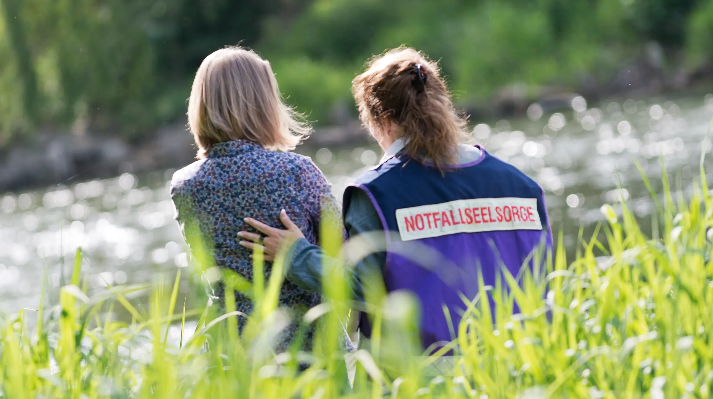 Eine Notfallseelsorgerin und eine Frau sitzen im Gras. Beide sind nur von hinten zu sehen. Die Notfallseelsorgerin legt der Frau die linke Hand auf den Rücken und möchte so die Frau beruhigen.