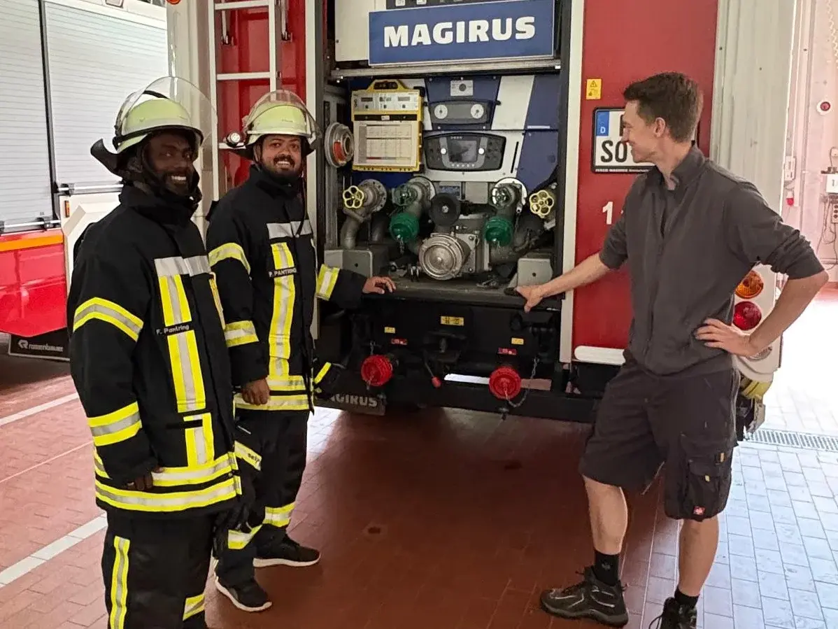 Zwei Männer in Feuerwehruniform und ein junger Mann in Shorts und T-Shirt stehen vor einem großen Feuerwehrfahrzeug, dessen hintere Seite geöffnet ist und den Blick auf die Gerätschaften im Innern freigibt.