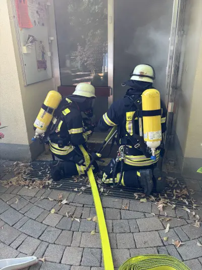 zwei Feuwehrleute in Schutzkleidung sitzen mit einem Schlauch in der Hocke vor der Eingangstür der Kita, es kommt Rauch aus der türe.