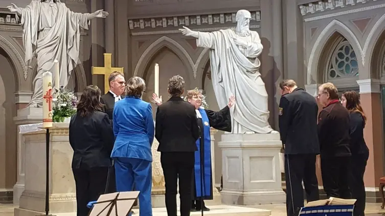 Die Pfarrerin spendet den Segen für einige Personen, die im Altarraum stehen. Im Hintergrund große Steinfiguren, die Jesus Christus und einen Apostel zeigen.