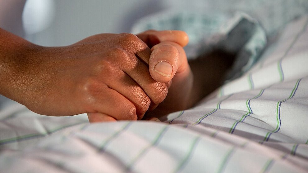 Eine Person liegt auf einem Bett. Ihre Hand wird von einer anderen Person gehalten. Auf dem Bild sind nur die Hände zu sehen.