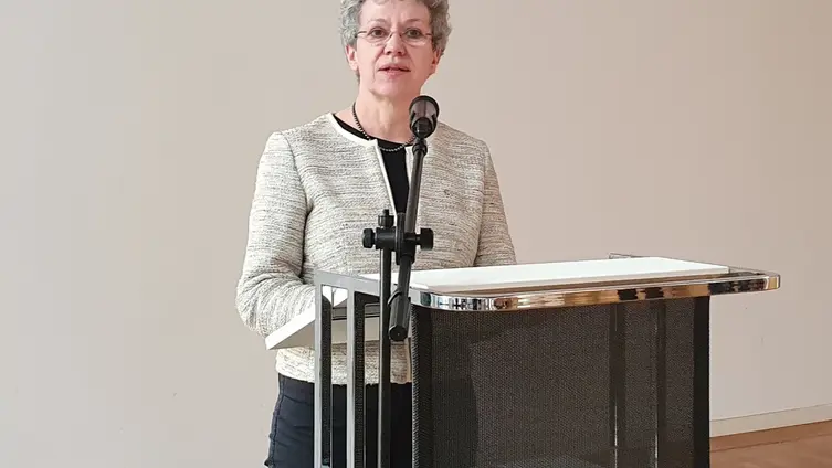 Frau Pfeiffer steht hinter dem Rednerpult vor einer weißen Wand.