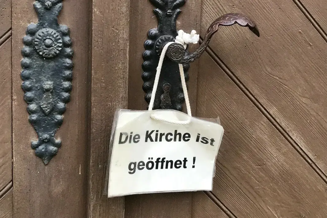 Weißes Schild hängt an Klinke der Kirchentür. Auf dem Schild steht: "Die Kirche ist geöffnet".