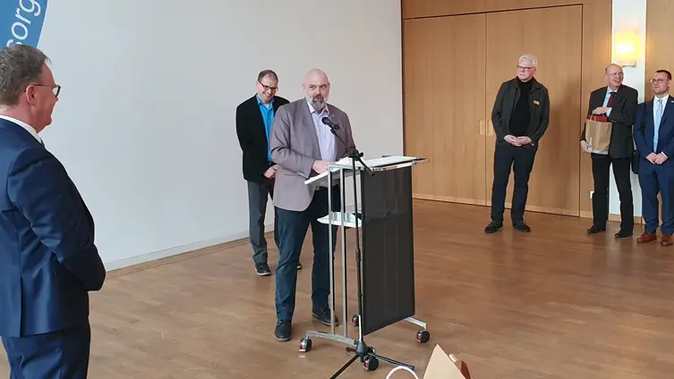 Ein Redner steht am Rednerpult, im Hintergrund weitere Gratulanten. 