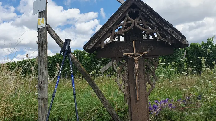 Neben einem Kreuz mit einer Jesusfigur stehen Wanderstöcke. Im Hintergrund wachsen Weinreben.