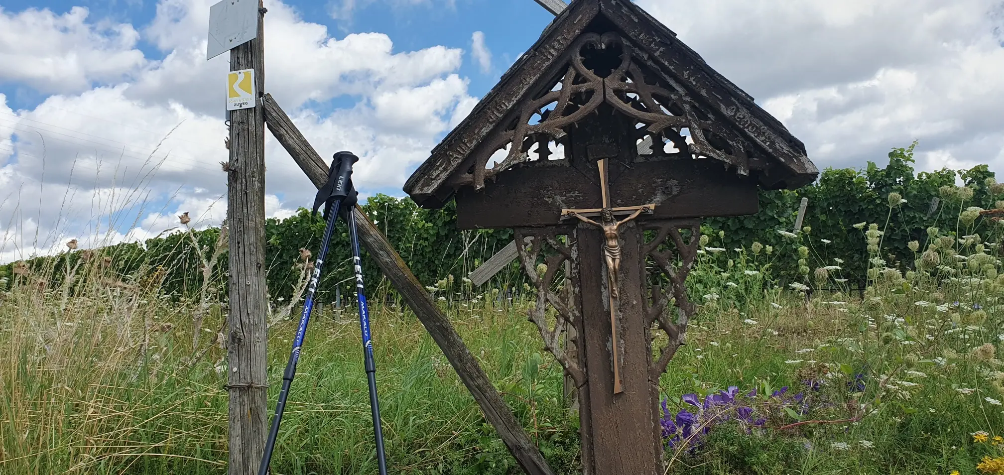 Neben einem Kreuz mit einer Jesusfigur stehen Wanderstöcke. Im Hintergrund wachsen Weinreben.
