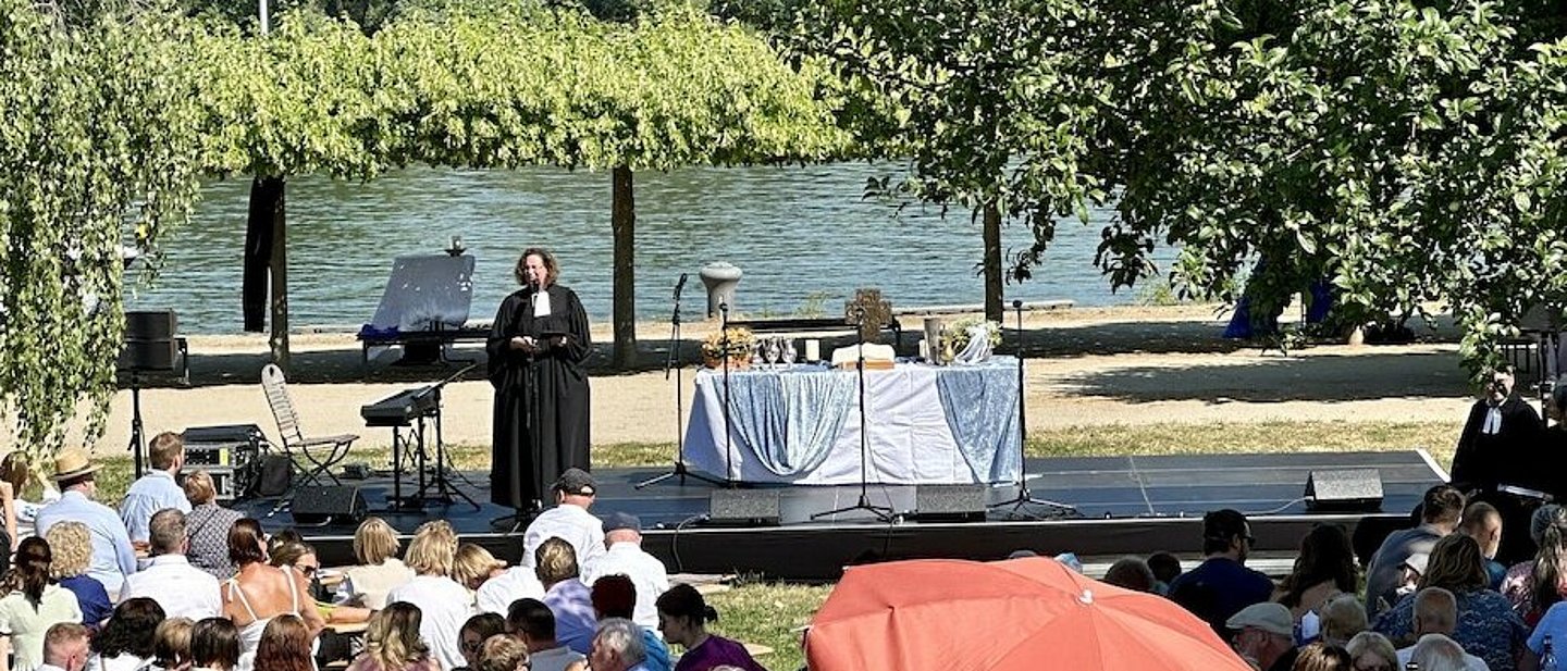 Tauffest in Ingelheim2023: Im Vordergrund sind die Besucher*innen im Bild, die auf Bänken vor Tischen sitzen. Dahinter steht eine kleine Bühne auf dem eine Pfarrerin steht und spricht. Daneben steht ein Altar mit diversen Taufutensilien.