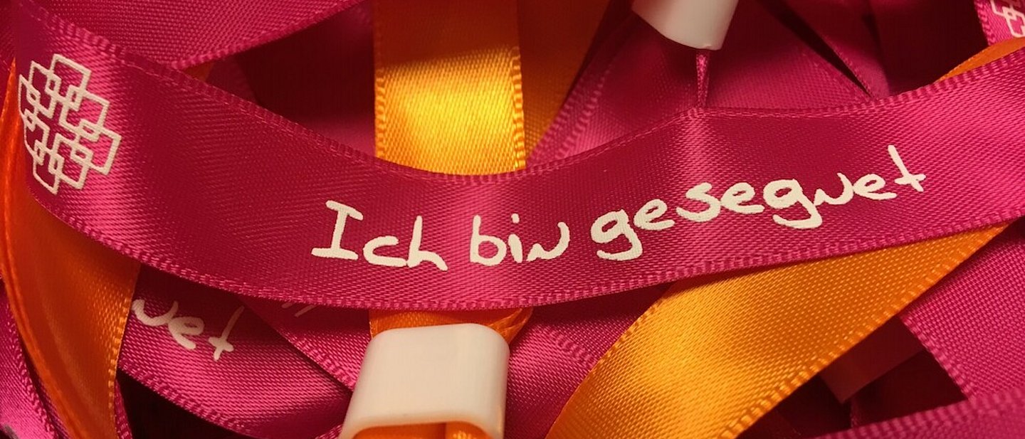Rote und orangene Stoffarmbänder mit der Aufschrift "Ich bin gesegnet" und dem Facettenkreuz.