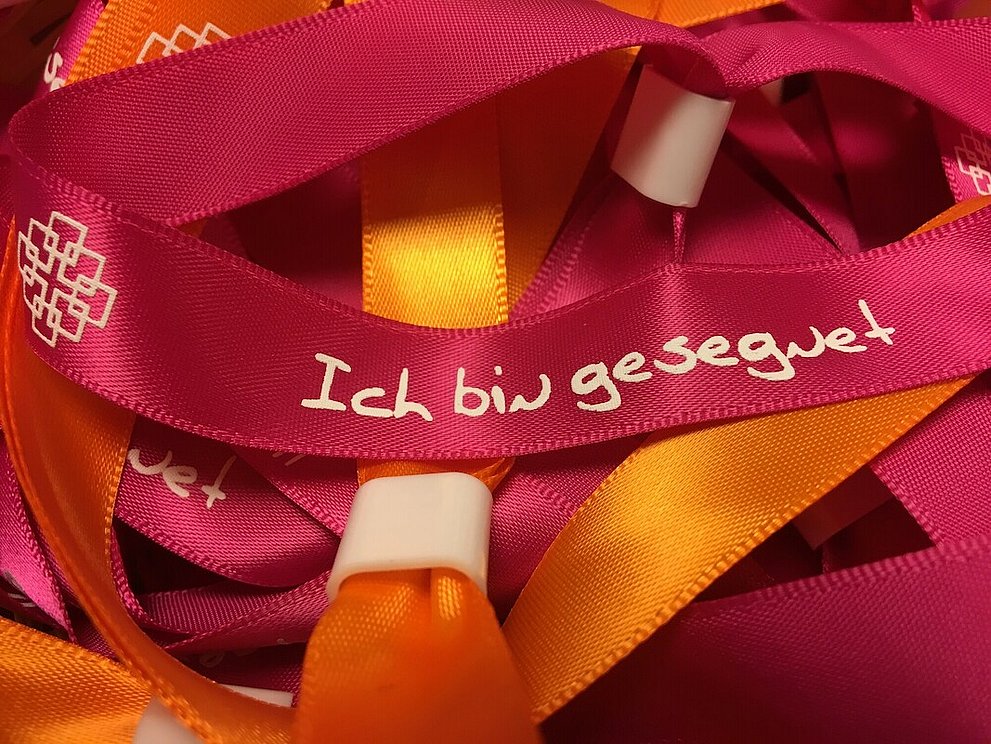 Rote und orangene Stoffarmbänder mit der Aufschrift "Ich bin gesegnet" und dem Facettenkreuz.