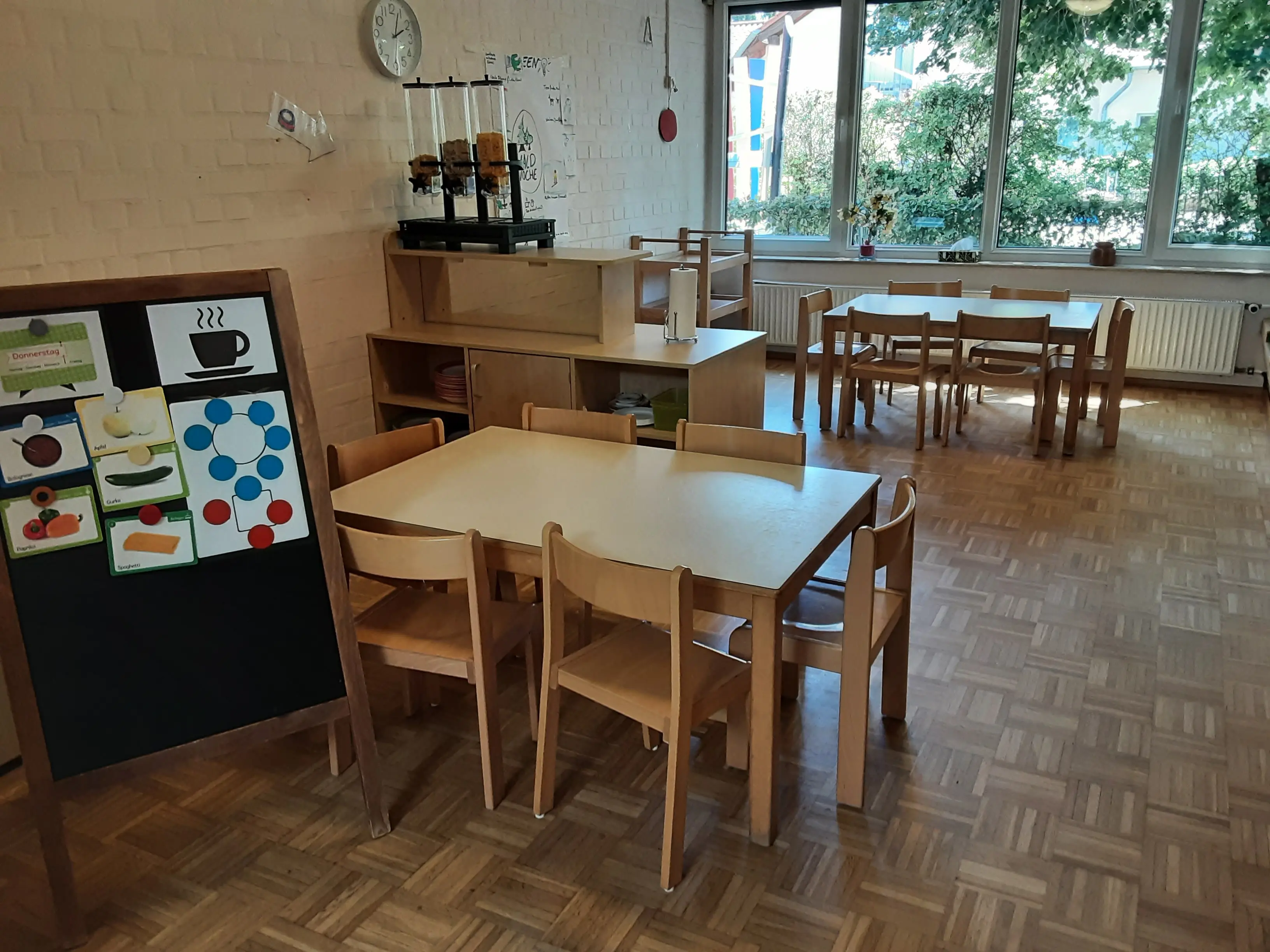 Cafeteria mit einem Tisch und Stühlenim Vordergrund, daneben eine Tafel mit dem Essenskarten. Im Hintergrund ein Buffet auf Kinderhöhe, dahinter noch ein Tisch und Fenster.