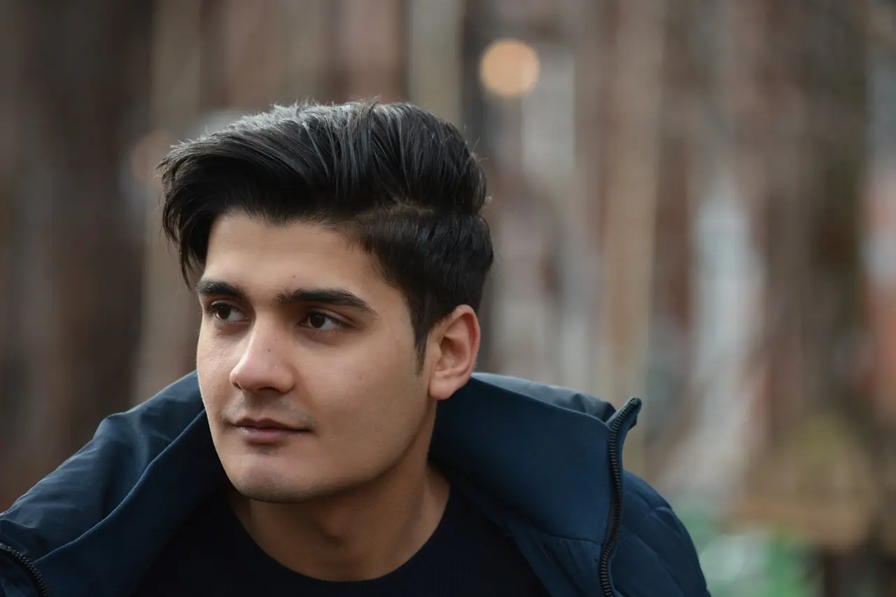 Das ist ein Nahaufnahmefoto eines jungen Mannes mit dunklem Haar und dunklen Augen. Er trägt ein dunkles T-Shirt und eine dunkle Jacke. Der Hintergrund ist unscharf und zeigt Bäume. Der junge Mann schaut leicht nach rechts weg von der Kamera.