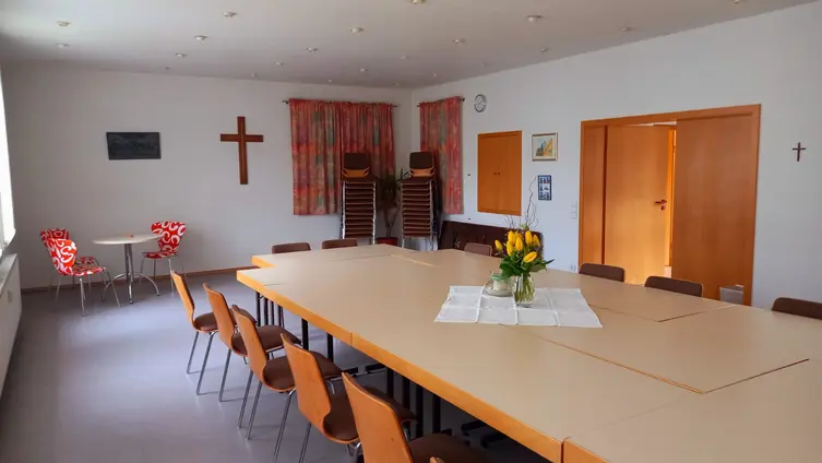 Ein heller Raum. In der Mitte steht ein großer Tisch, um den Tisch herum stehen Stühle. An der Wand hängt ein Kreuz